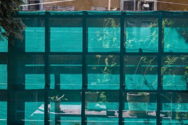 Bahar güneşli bir günde aydınlanan yeşil örgü naylon kumaş tel örgü. Arkadaki saksılarda bitkilerin silueti. Arka planda eski bir evin duvarı. Bratislava, Slovakya.