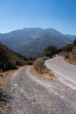 Arka planda yüksek bir dağ olan Girit 'in merkezi. Taş toprak, ağaç ve çalılarla kaplı bir yol ve kıvrım. Parlak mavi gökyüzü. Girit, Yunanistan.