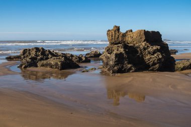 Atlantik Okyanusu kıyısında kum ve kayalarla. Okyanus ufku ve parlak mavi bir gökyüzü. Vahşi dalgaların havasındaki sis. Essaouira, Fas.