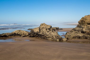 Atlantik Okyanusu kıyısında kum ve kayalarla. Okyanus ufku ve parlak mavi bir gökyüzü. Vahşi dalgaların havasındaki sis. Essaouira, Fas.