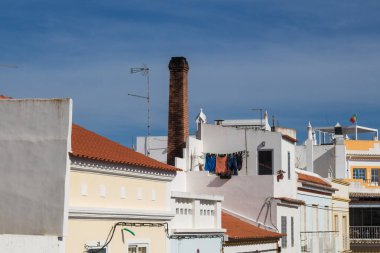Çoğunlukla turuncu ya da düz çatılı evlerin beyaz cepheleri. İkisinin arasında baca var. Mavi gökyüzünde hafif bulutlar. Alvor, Algarve, Portekiz.