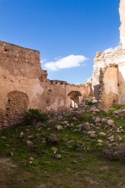 Terk edilmiş evin kalıntılarında cepheli olmayan duvarlar. Bahçede yeşil çimenler ve yıkılmış duvarlardan bir sürü taş var. Beyaz bulutlu parlak mavi gökyüzü. Dar Caid Hacı (aynı zamanda Hoca olarak da bilinir), Fas.