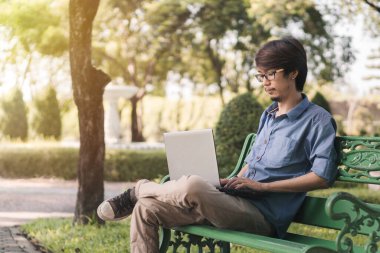 Genç Asyalı bir adam laptopuyla parktaki bankta çalışıyor. Tatil zamanı..