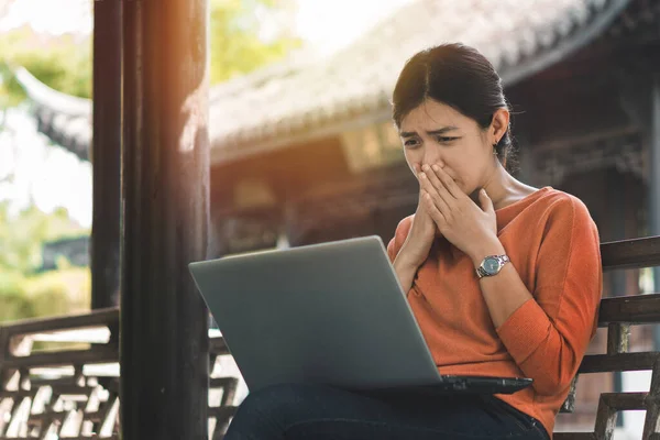 Genç Asyalı kadın dizüstü bilgisayarıyla çalışıyor ve tatilde parkın dışında kendini endişeli hissediyor..