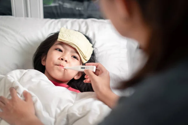 Asyalı anne sabah saatlerinde yatakta sıcaklık ölçer, hasta çocuk yüksek ateşi, seçici odağı, sağlıklı ve enfeksiyon konseptini azaltmak için havalı havluya sahiptir.