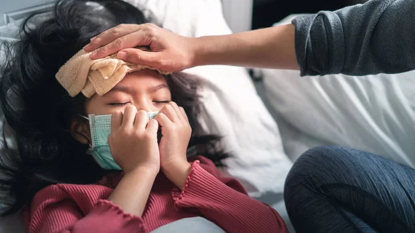 アジアの母親は 病気の少女額に高熱を減らすためのクールなタオルを取ります 女の子は医療保護顔マスクと咳を身に着けています 選択的な焦点 健康と感染の概念 — ストック写真