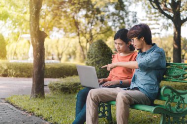Genç Asyalı bir adam ve kadın dizüstü bilgisayarlarıyla çalışıyor ve tatil zamanlarında parktaki bir bankta monitörü işaret ediyorlar..