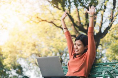 Mutlu genç Asyalı kadın dizüstü bilgisayarıyla çalışırken gülümseyecek ve tatil zamanı parktaki bir bankta el ele verecek..