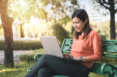 Mutlu genç Asyalı kadın laptopuyla çalışıyor. Parktaki bankta tatilde..