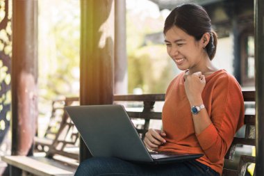 Genç Asyalı mutlu bir kadın dizüstü bilgisayarıyla çalışıyor ve tatilde açık havada parkta online galibiyetinin tadını çıkarıyor..