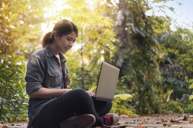 Yaz tatili konsepti, mutlu Asyalı kadın dizüstü bilgisayarıyla gülümseyecek ve tatilinde tropik ormanda oturacak..
