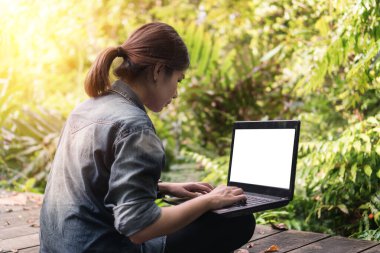 Yaz tatili konsepti, mutlu Asyalı kadın dizüstü bilgisayarıyla gülümseyecek ve tatilinde tropikal ormanda oturacak, mesajınızı kırpma yolu ile ayarlamak için beyaz ekran dizüstü bilgisayarı.