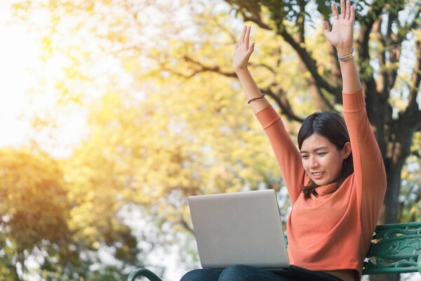 Счастливая молодая азиатская женщина будет улыбаться, работая со своим ноутбуком и руки вверх на скамейке в парке на открытом воздухе во время отпуска
.