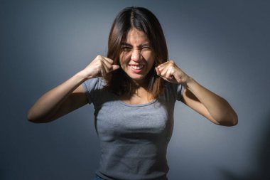 Duygusal konsept, kızgın, sevimli Asyalı kadının portresi gri elbiseli kameraya bakıyor ve koyu mavi arka planda yumruğunu gösteriyor..