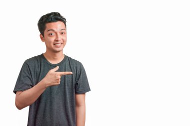 Gülümseyen mutlu Asyalı genç adam gri tişört giyer ve beyaz arka planda izole edilmiş, kırpma yolu olan fotokopi alanını gösterir..