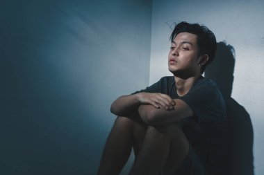 Genç bunalımlı Asyalı adam karanlık odada yalnız başına oturuyor. Düşük ışık ortamı, seçici odaklanma, travma sonrası stres bozukluğu zihinsel sağlık konsepti..