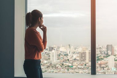Depresyonda olan Asyalı genç bir kadın akşam vakti pencerenin kenarında dikiliyor. Düşük aydınlık bir çevresi, travma sonrası stres bozukluğu, zihinsel sağlık konsepti, seçici odaklanma.