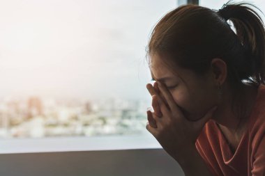 Genç depresif Asyalı kadın akşam vakti pencerenin kenarında yalnız başına oturuyor. Düşük ışık ortamı, travma sonrası stres bozukluğu, zihinsel sağlık, seçici odaklanma....