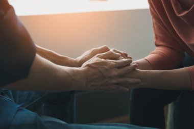 Psikolog, pencerenin yanında düşük ışık ortamı, travma sonrası stres bozukluğu, zihinsel sağlık konsepti ve seçici odaklanma için depresyonda olan Asyalı genç bir kadınla el ele tutuşuyor..