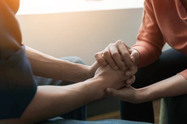 Travma sonrası stres bozukluğu, zihinsel sağlık konsepti, psikolog oturuyor ve pencerenin yanında düşük aydınlık çevreye sahip genç Asyalı adamı cesaretlendirmek için el ele tutuşuyor..