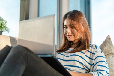 Genç Asyalı kadın dizüstü bilgisayarda çalışıyor ve evdeki kanepede oturuyor..