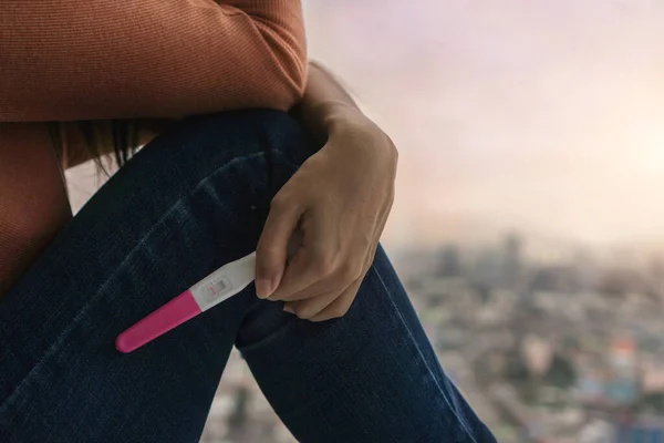 Unhappy young asian woman holding pregnancy test showing a negative result and sit down near big window city view on evening time, Wellness and healthy concept, Infertility problem, Selective focus.