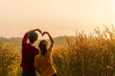 Asyalı kadın ve erkek ayakta duruyor ve aşk pozu veriyor. Çayırda birlikte imzalıyorlar ve gün doğumunda uzaklara bakıyorlar..