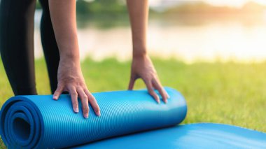 Asyalı genç bir kadın mavi yoga minderi ya da spor minderi yuvarlıyor. Park, sağlıklı konsept ve zihin-vücut geliştirme konseptinde egzersiz yapmaya hazır olun..