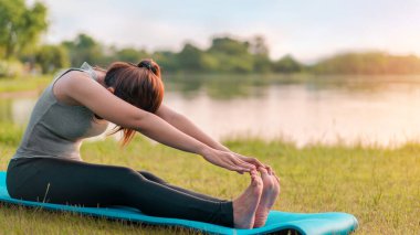 Genç Asyalı kadın egzersiz kıyafeti giyiyor ve parktaki spor minderinde yoga yapmadan önce geriniyor, sağlıklı konsept, zihin-vücut geliştirme konsepti.;