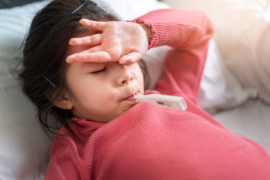 Asyalı bir kız sabah yatağında ağzında dijital termometreyle kendini ve alnını ölçüyor, yüksek ateş seçici odaklı hasta çocuk, sağlıklı ve enfeksiyon konsepti