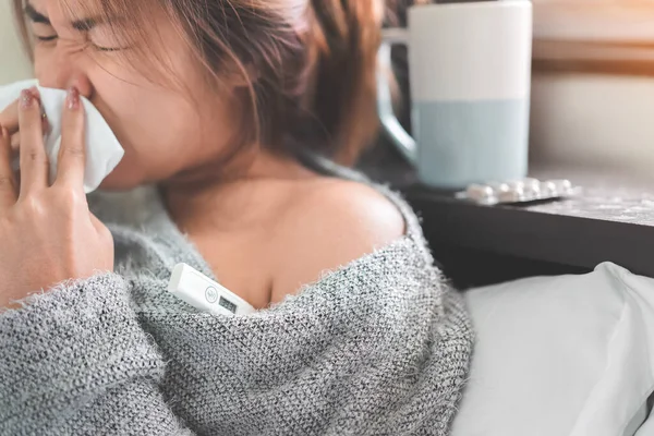 Sick asian woman have hight fever flu sneezing into tissue and checking temperature by digital thermometer on bed in bedroom, Medical and health concept, Selective focus.