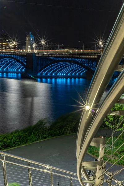 Handrail shining in foreground with blue lights glowing under bridge at night.  (Charles River, Boston, Massachusetts, USA)