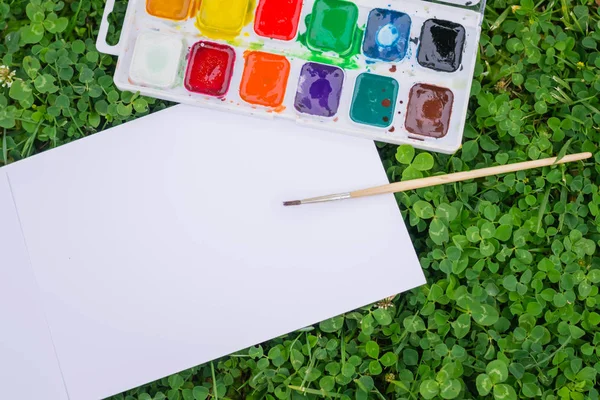 Çimlerin üzerinde çok renkli suluboya boyalar bulunan bir palet. Bir kağıt parçasının yanında, bir çizim albümü. Bir şeyler yazabileceğin bir yer var. Kağıtta farklı boya fırçaları var..