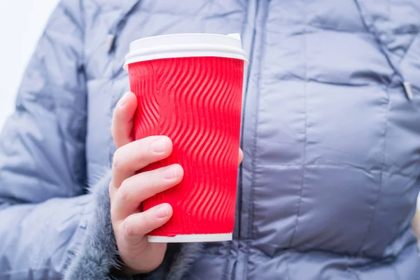 Elinde sıcak içecekle kağıt bardak tutan genç bir kadın. Beyaz plastik kapaklı kırmızı bardak. Soğuk havada açık havada ısınmak için. Yeni yıl ve Noel tatili. Kış Yolculuğu.