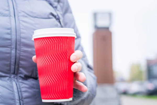 Elinde sıcak içecekle kağıt bardak tutan genç bir kadın. Beyaz plastik kapaklı kırmızı bardak. Soğuk havada açık havada ısınmak için. Yeni yıl ve Noel tatili. Kış Yolculuğu. Saat arka planda. Kahve zamanı.