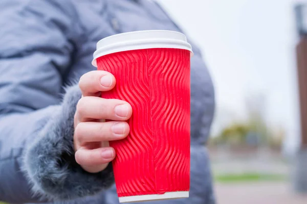 Elinde sıcak içecekle kağıt bardak tutan genç bir kadın. Beyaz plastik kapaklı kırmızı bardak. Soğuk havada açık havada ısınmak için. Yeni yıl ve Noel tatili. Kış Yolculuğu. Saat arka planda. Kahve zamanı.