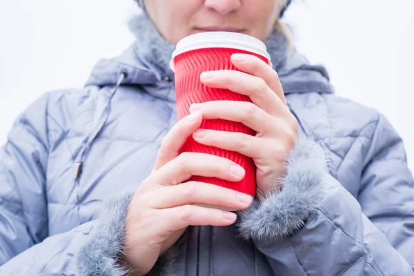 Red paper cup of coffee in the hands of a woman. Young blond woman dressed in a winter gray down jacket. On the head is a blue hat. She drinks coffee on the street. It is heated by coffee.
