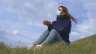 Yeşil tepede oturan tıbbi maskeli genç kadın.