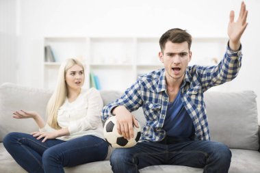 Kızgın adam bir futbol topu izler ve kanepedeki kadının yanında el kol hareketi yapar.