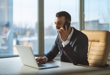 İş adamı masada dizüstü bilgisayarla telefon ediyor.