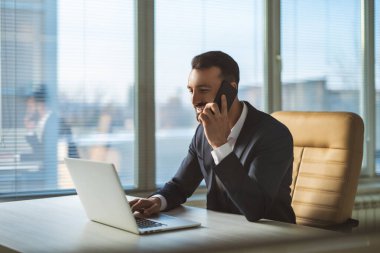 İş adamı masada dizüstü bilgisayarla telefon ediyor.