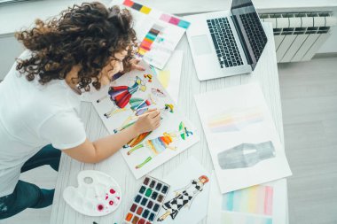 Kıvırcık kadın masada dizüstü bilgisayarla resim yapıyor. Yukarıdan görünüm