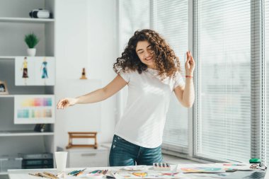 Kıvırcık sanatçı masanın yanında resimlerle müzik dinliyor.