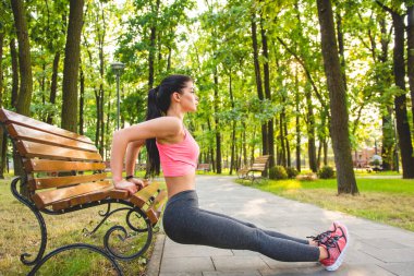 Sporcu kadın parkta bir bankla egzersiz yapıyor.