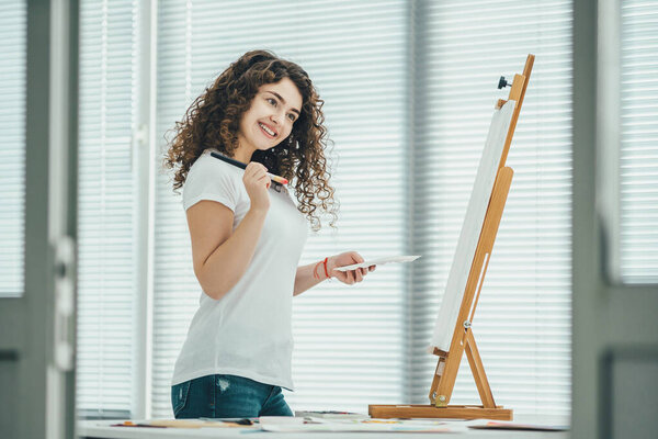 Счастливая кудрявая женщина рисует картину на мольберте
