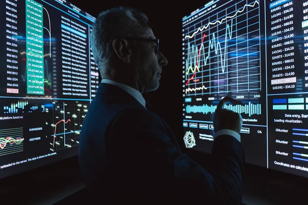 İş adamı karanlık odadaki büyük dijital ekranın yanında duruyor.