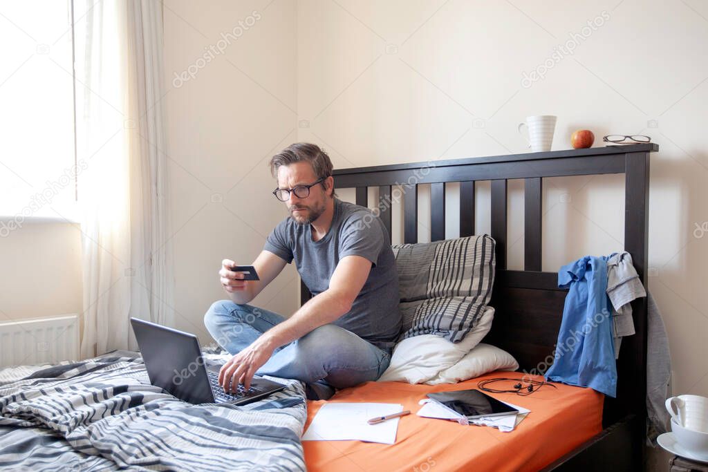 Man making online payment, sitting on bed whilst working from home