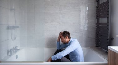Depresyondaki adam banyoda oturuyor, tamamen giyinik, duş açık. 