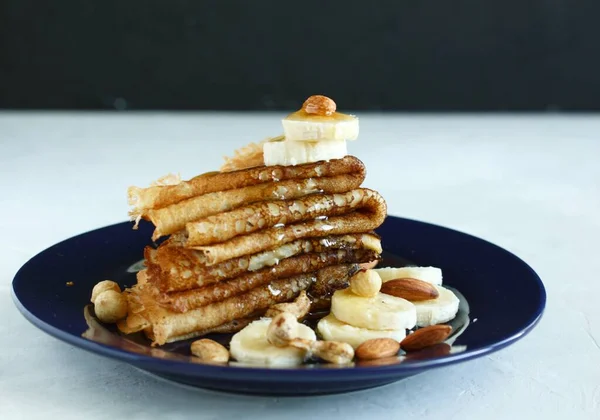A stack of pancakes with nuts and a banana and liquid golden honey. Liquid honey flows through pancakes. On a dark blue plate