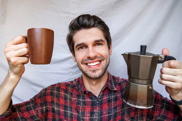 Young handsome man with a wide open smile holding a cup of coffee and a moka pot in a shirt